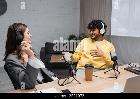 Fröhliches Multiethnisches Radio mit Kopfhörern, die in der Nähe professioneller Mikrofone, Kaffee zum Mitnehmen, Notebooks und Smartphones auf dem Tisch in modernen Breiten sprechen Stockfoto