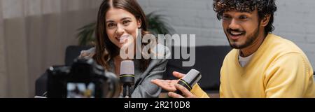 Bärtiger und lockiger indischer Podcaster in gelbem Pullover, der sich im Mikrofon unterhält, in der Nähe einer lächelnden, brünetten Kollegin, die im Radio-Studio einen Blazer trägt, während sie sich erholt Stockfoto