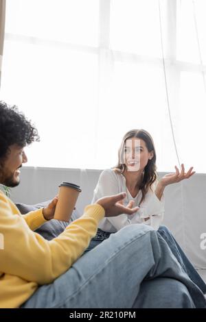 Ein geschwungener indischer Mann in gelbem Pullover und Jeans, der Kaffee zum Mitnehmen hält, während er sich mit einer positiven brünetten Kollegin in weißer Bluse auf s unterhält Stockfoto