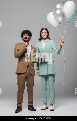 Ein lächelnder junger indischer Veranstalter, der mit einem Mikrofon sprach, während er neben einem brünetten Kollegen stand, mit bunten Ballons auf grauem Hintergrund. Stockfoto