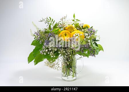 Blumenstrauß aus Frühlingsblumen in Gelb, Blau und Grün in einer Glaskanne vor hellgrauem Hintergrund mit Kopierbereich, Weihnachtsgrußkarte ausgewählt Stockfoto