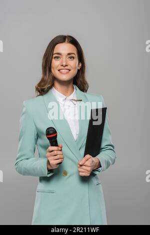 Porträt von positiver Brünette Moderator des Ereignisses in blauem Anzug hält Mikrofon und Clipboard während der Party, während er in die Kamera schaut und isoliert steht Stockfoto