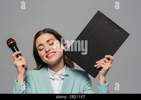 Lächelnder brauner Veranstaltungsort mit geschlossenen Augen in blauer Jacke, Mikrofon und Klemmbrett während der Feiertagsfeier isoliert auf grauem Stockbild Stockfoto