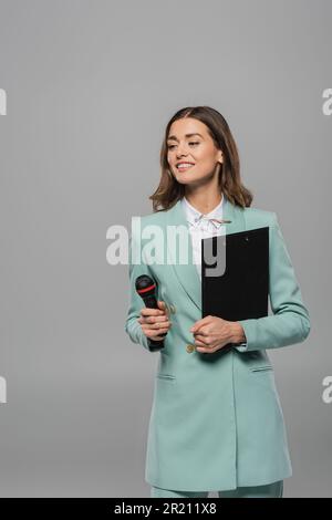 Fröhliche Brünette, Veranstaltungsort in blauer Jacke und Hemd mit Mikrofon und Klemmbrett, während die Feier isoliert auf grauem Stock abgewandt wird Stockfoto