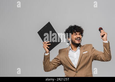 Aufgeregter junger indischer Veranstalter mit lockigem Haar in formeller Kleidung Augen schließen, während der Vorstellung Klemmbrett und Mikrofon in der Hand gehalten werden, isoliert auf grau Stockfoto