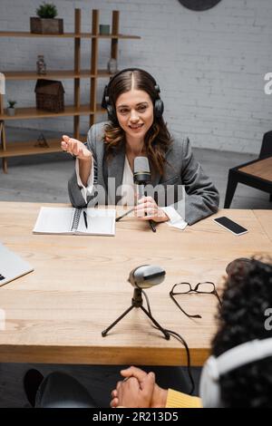 Lächelnder und charmanter Ansager in Kopfhörern, der in der Nähe von Mikrofon, Smartphone und Notebook mit Stift spricht und das Interview mit Bl aufnimmt Stockfoto