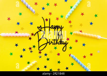 Die Inschrift Happy Birthday auf gelbem Hintergrund. Festlicher Hintergrund mit Kerzen, Konfetti in Form von Sternen mit Kopierraum Stockfoto