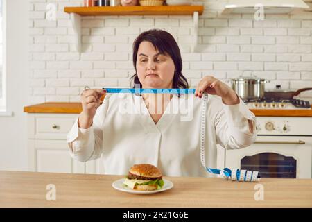 Eine fette, übergewichtige junge Frau, die in der Küche sitzt und ein Maßband mit einem Burger auf dem Tisch hält. Stockfoto