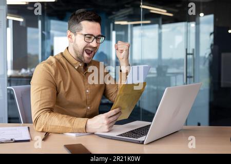 Ein junger männlicher Student, der im Büro studiert, an einem Laptop mitarbeitet und einen Brief von Yakits liest, den er per Post erhalten hat. Freut sich über die gute Nachricht, IS Stockfoto