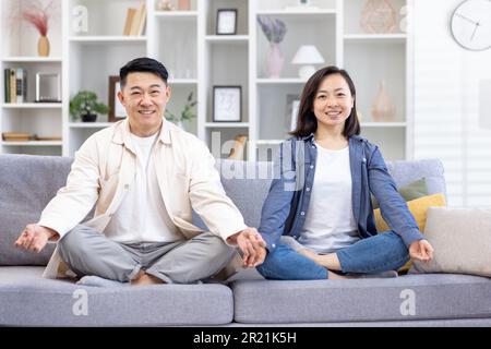 Porträt einer jungen asiatischen Familie, ein Paar, das zu Hause Yoga macht. Ein Mann und eine Frau sitzen auf einem Sofa in Lotusposition und lächeln in die Kamera. Kümmern Sie sich um ihre psychische Gesundheit. Stockfoto