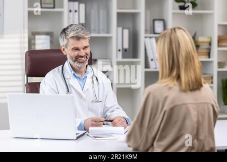 Arzttermin. Ein erfahrener Arzt sitzt im Büro an einem Tisch mit einem Laptop und empfängt Patienten, konsultiert und spricht mit einer jungen Frau. Stockfoto