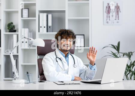 Lächelnder junger hispanischer Doktor Student, Praktikant, Assistentin sitzt im Büro am Laptop, studiert, übt, besteht Prüfung. Begrüßt mit seiner Hand, stellt sich der Kamera vor. Stockfoto