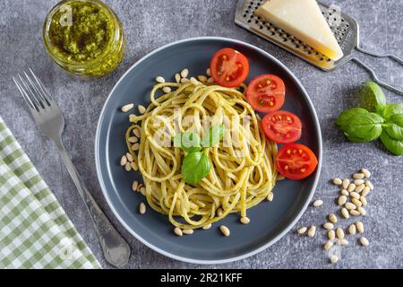 Nudelspaghetti mit Pesto-Sauce und frischen Basilikumblättern in schwarzer Schüssel auf grauem Betonhintergrund, serviert mit Zutaten. Traditionelles italienisches Cu Stockfoto