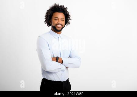 Ein hübscher, geschwungener Mann, der in die Kamera schaut, mit gekreuzten Armen, isoliert auf weißem Hintergrund. Afroamerikanischer männlicher Angestellter mit lockigem Haar in einem formellen weißen Hemd mit gefalteten Händen Stockfoto