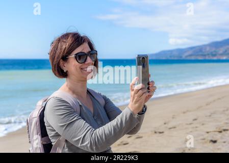 Lächelnde Touristin, die an einem sonnigen Herbsttag an einem Sandstrand in Kalifornien mit einem Smartphone ein Foto macht Stockfoto