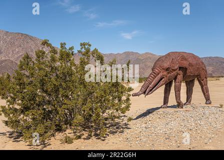 Borrego Springs, CA, USA - 24. April 2023: Braune rostige prähistorische Riesenantiquen-Mammuts-Statue auf sandigem Wüstenboden mit grüner Buschunde Stockfoto