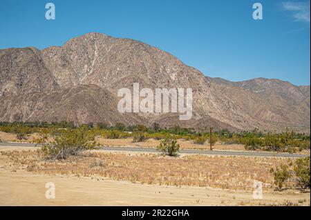 Borrego Springs, CA, USA - 24. April 2023: Asphaltstraße durch eine breite Sandwüstenlandschaft mit grünen Büschen unter blauer Wolkenlandschaft. Beige-graue Berge Stockfoto