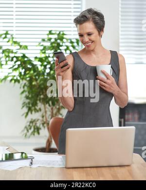 Immer in Kontakt. Eine reife Geschäftsfrau, die ihr Handy durchsucht, während sie im Büro Kaffee trinkt. Stockfoto