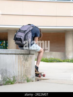 College-Student, der sich mit seinem elektrischen Skateboard und Rucksack beugte, Campus, und zwischen den Klassen auf sein Telefon schaute. Stockfoto