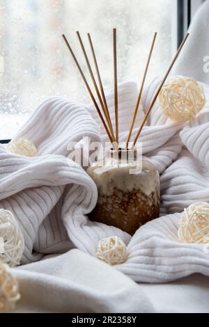 Aroma-Reed-Diffusor. Flaschenbehälter mit Holzstäbchen diffussiert ätherische Öle, um Ihr Heim Duft Konzept Herbsturlaub in gemütlichen Heim auf dem Fenstersill Hygge Ästhetische Atmosphäre auf gestricktem weißen Pullover. Stillleben im Mikromoment, aufrichtiges langsames Leben. Psychische Gesundheit Wohlbefinden Übungen regnen draußen Stockfoto