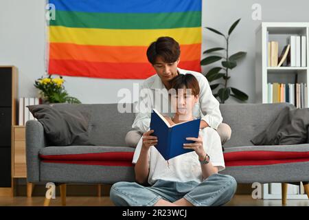 Glückliches schwules Paar, das zu Hause ein Buch auf dem Sofa liest und am Wochenende Zeit miteinander verbringt. LGBTQ-Menschen, Lebensstil und Liebesgefühle Stockfoto