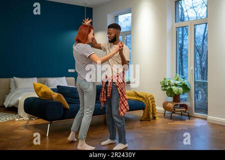 Liebevolles junges, vielfältiges Paar, das romantischen Tanz im modernen Schlafzimmer mit Panoramafenstern tanzt. Stockfoto