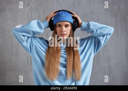 Ein junges, hübsches, langhaariges Mädchen mit blauem Pullover und blauer Mütze trägt DJ-Kopfhörer auf dem Kopf und hält sie mit den Händen. Musik und Schönheit Stockfoto