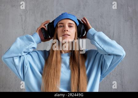 Ein junges, hübsches, langhaariges Mädchen mit blauem Pullover und blauer Mütze trägt DJ-Kopfhörer auf dem Kopf und hält sie mit den Händen. Musik und Schönheit Stockfoto