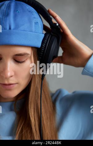 Nahaufnahme eines jungen, hübschen, langhaarigen Mädchens mit blauem Pullover und blauer Mütze trägt DJ-Kopfhörer auf dem Kopf und hält sie mit den Händen. Musik und Stockfoto