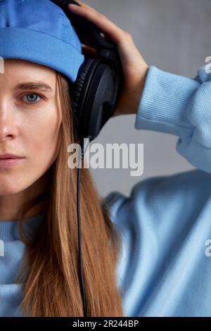 Nahaufnahme eines jungen, hübschen, langhaarigen Mädchens mit blauem Pullover und blauer Mütze trägt DJ-Kopfhörer auf dem Kopf und hält sie mit den Händen. Musik und Stockfoto