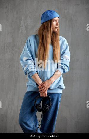 Porträt eines jungen, hübschen, langhaarigen Mädchens mit blauem Pullover, Hut und Jeans, das DJ-Kopfhörer in der Hand hält. Musik, Spaß und Schönheit Stockfoto
