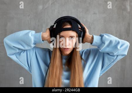 Ein junges, hübsches, langhaariges Mädchen mit blauem Pullover und blauer Mütze trägt DJ-Kopfhörer auf dem Kopf und hält sie mit den Händen. Musik und Schönheit Stockfoto