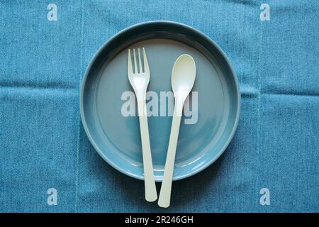 Leerer marineblauer Keramikteller mit Gabel und Löffel auf blauem Textiltischhintergrund. Draufsicht Stockfoto