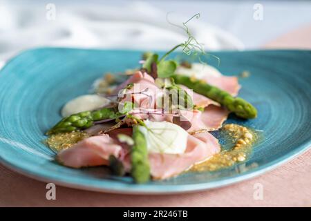 Selektiver Fokus einer Vorspeise mit Spargel, Taleggio, Erdnüssen und Rosmarinschinken auf einem blauen Teller. Pinkfarbenes Tuch und weiße Serviette auf dem Tisch. Stockfoto