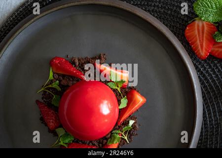 Selektiver Fokus von Erdbeeren auf Brownie Crumbs. Kugelförmiger Quark mit glatten Oberflächen und Spiegelglasur. Rotes Dessert auf dem schwarzen Teller. Stockfoto