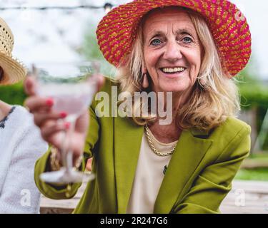 Porträt einer langhaarigen blonden älteren Frau mit rotem Strohhut, die mit Freunden am Tisch sitzt, lächelt und einen Toast auf die Kamera ausspricht Stockfoto