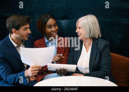 Vielseitige, begeisterte Geschäftspartner in formellen Anzügen sitzen mit Unterlagen am Tisch und diskutieren Projektdetails in einem modernen Arbeitsplatz Stockfoto