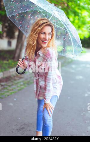 Fröhliche junge rothaarige Großstadtmodel-Frau mit langen welligen Haaren und transparentem Regenschirm, die draußen posiert Stockfoto