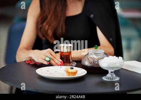 Das Mädchen genießt eine Tasse türkischen Tee allein in einem gemütlichen Café. Die Hände schließen sich mit Glas und Teekanne auf dem schwarzen Tisch. Stockfoto