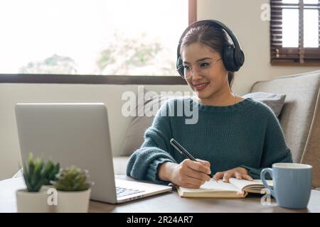 Frau, die sich Notizen im Notebook macht, während sie zu Hause ein Notebook im Wohnzimmer verwendet. Fokussierte Frau, die in Notizblock schreibt Stockfoto