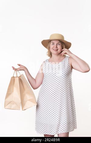 Weibliche Käuferin in weißem Kleid mit schwarzen Punkten und Strohhut, die zwei handwerklich gefertigte Einkaufstaschen in einer Hand hält Stockfoto