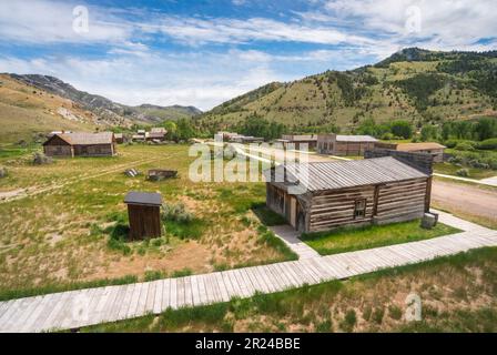 Eine verwitterte Blockhütte und ein kleines Plumpsklo stehen neben einem Plankensteg, mit der Geisterstadt Bannack State Park unter grünen Hügeln, Montana. Stockfoto