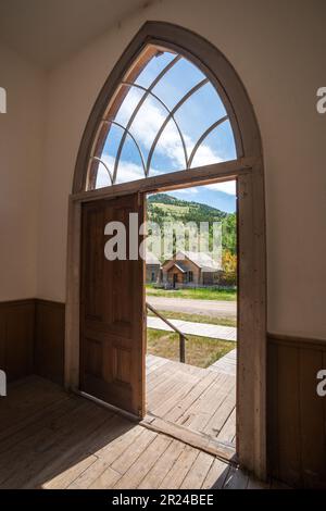Ein hoher spitzbogiger Eingang mit einem gewölbten Maßwerkfenster darüber steht offen zu einer Bretterveranda und Geisterstadtgebäuden im Bannack State Park, Montana. Stockfoto