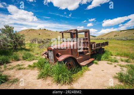 Ein verrosteter Vintage-Truck mit einem Holzbett steht im Sand und grünen Gras, mit Blockhäusern im Hintergrund, im Bannack State Park, Montana. Stockfoto