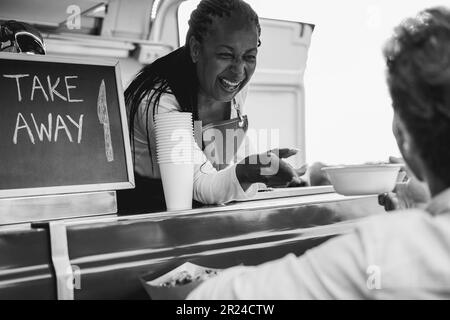 Afrikanische Seniorin, die Essen zum Mitnehmen im Food Truck serviert - weicher Fokus auf Chefgesicht - Schwarz-Weiß-Schnitt Stockfoto