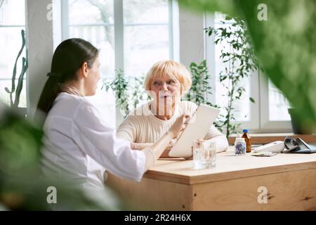 Seniorin beim Arztbesuch, sitzt am Tisch im Büro, hat Termin und Beratung. Krankenschwester zeigt Informationen auf dem Tablet an Stockfoto
