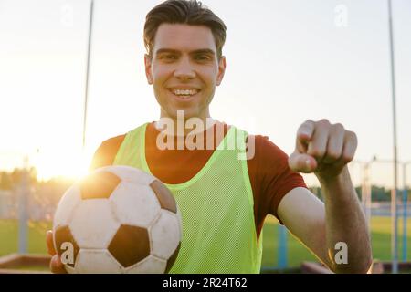 Ein junger, lächelnder Fußballspieler, der den Fußball hält und mit dem Finger auf die Kamera und auf dich zeigt Stockfoto