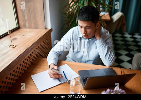 Junger, attraktiver männlicher Freiberufler, Student sitzt am Fenster im Innern des Cafés der Stadt und arbeitet an Laptops und Papieren. Produkteigentümer erstellt neu Stockfoto