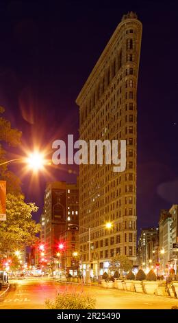 Das Flatiron Building leuchtet warm unter Straßenlaternen unter einem indigo-Himmel vor der Morgendämmerung. Der schwache Verkehr hinterlässt leichte Wege bei zeitlicher Belichtung. Stockfoto