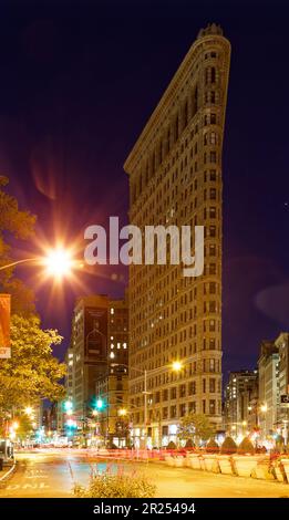 Das Flatiron Building leuchtet warm unter Straßenlaternen unter einem indigo-Himmel vor der Morgendämmerung. Der schwache Verkehr hinterlässt leichte Wege bei zeitlicher Belichtung. Stockfoto